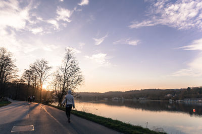 Rear view of woman walking on road by lake against sky during sunset