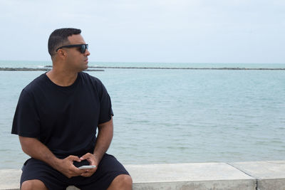 Man wearing sunglasses using mobile phone by sea against sky