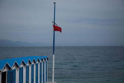 Flag pole by beach huts at riverbank against sky