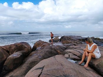 People sitting on rocks by sea against sky