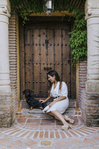 Smiling woman playing with dog while sitting at house doorway