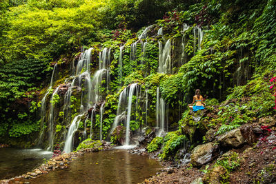 Rear view of woman practicing yoga while sitting on rock against waterfall in forest