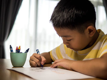 Surface level of boy drawing on table while sitting against window at home