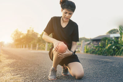 Full length of young woman suffering from knee pain while crouching on road