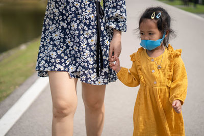 Little girl holding mother's hand wearing healthy face mask walking on the road.