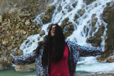 Young woman standing with arms outstretched against waterfall