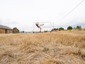 Man hanging from soccer goal on grassy field against sky