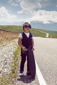 Fashionable child boy in white cap sneakers and vest stands with a skate, on road  mountain everest