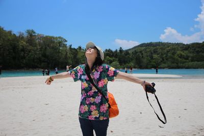 Woman with arms outstretched holding camera while standing against sea at beach