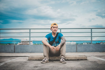 Portrait of young man sitting on railing against sky