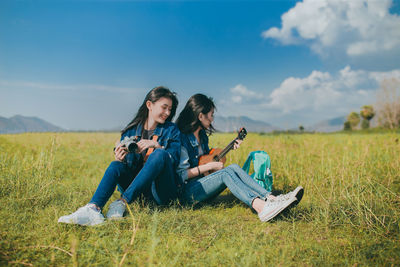 Young women sitting on grass while holding ukulele and camera against sky