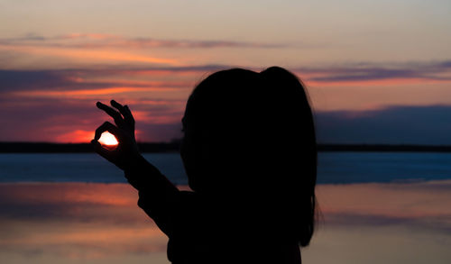 Silhouette woman showing ok sign against sky during sunset