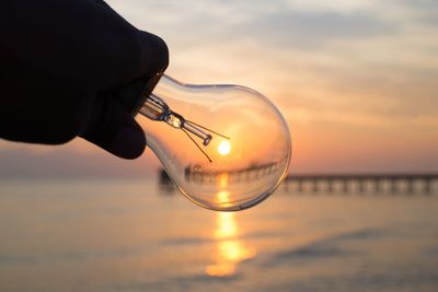 Close-up of hand holding light bulb against sea during sunset