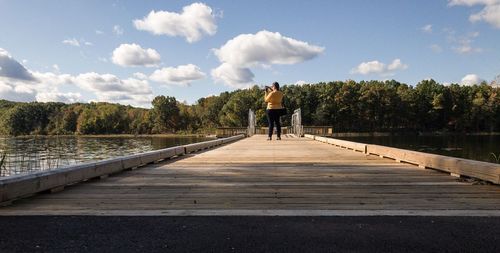 Rear view of mid adult woman photographing while standing on footbridge over river against sky