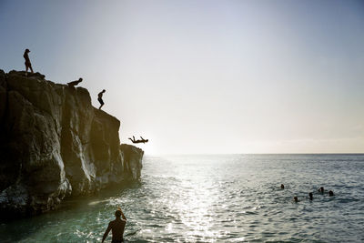 Friends jumping from cliff to sea against sky