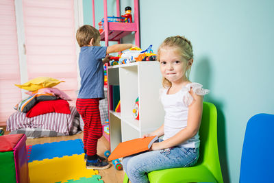 Portrait of girl sitting on chair by brother with toys over shelf