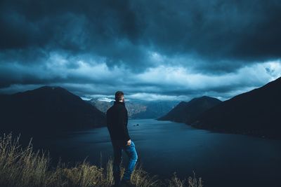 Rear view of man standing on mountain by river against cloudy sky