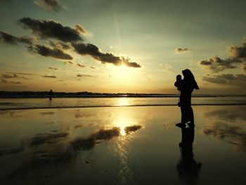 Silhouette woman with child standing on beach against sky during sunset