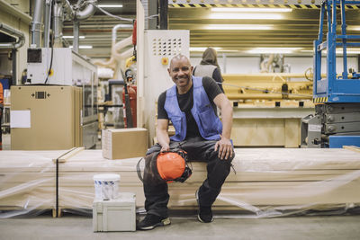 Portrait of happy carpenter sitting with hardhat in warehouse