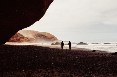 Rear view of men standing on shore at beach against sky