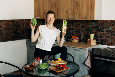 Cheerful young woman preparing green salad at home and jokingly throwing vegetables up