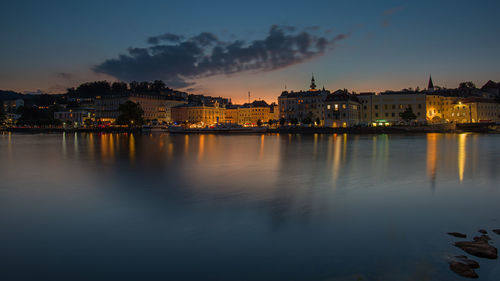 Illuminated city by lake against sky during sunset