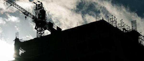 Low angle view of silhouette crane by building against sky