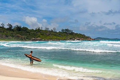 Man wearing swim trunks, standing on paradise beach, going surfing in turquoise ocean with waves