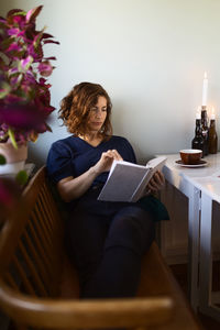 Adult female reading interesting book while sitting at table decorated with burning candles at home