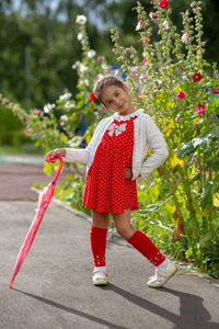 A cute little girl in a red dress and a white blouse with a pink cane umbrella stands on a summer 