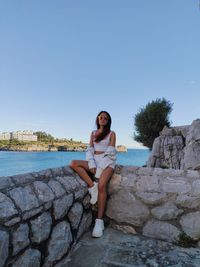 Portrait of young woman sitting on shore against clear sky