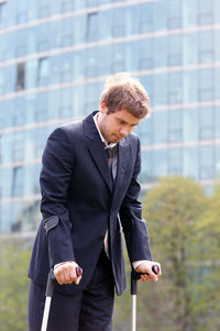 Businessman walks through a park in the city with crutches after knee injury