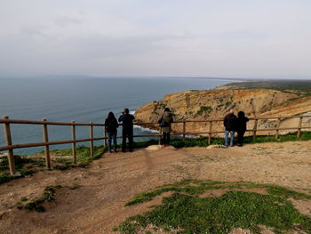 Rear view of people standing by railing on mountain by sea against sky