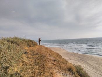 Man standing on grassy hill at beach against cloudy sky