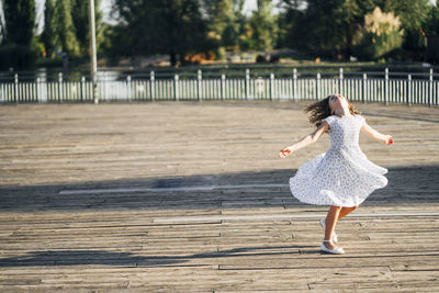 Cute teen girl spinning with hands apart on boardwalk of embankment