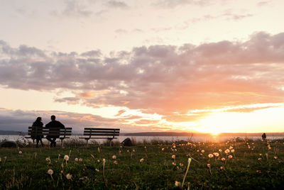 Couple sitting on bench against cloudy sky during sunset