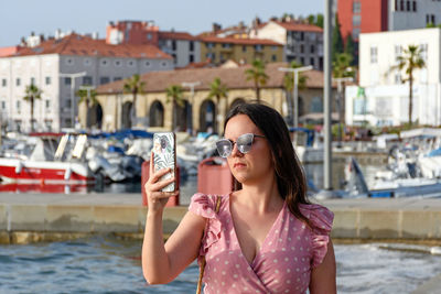 Beautiful young woman wearing pink dress, using mobile phone, taking photos on waterfront in city