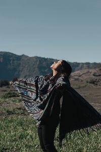 Smiling woman with arms outstretched standing on land against clear sky