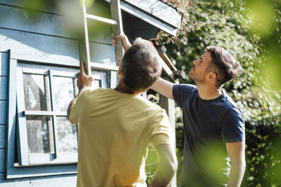 Father and son holding ladder by house during sunny day