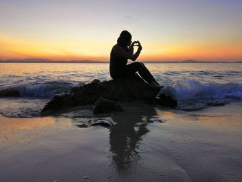 Man sitting on shore at beach against sky during sunset