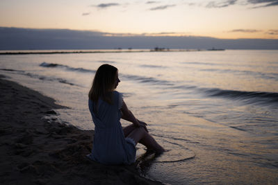 Woman sitting on shore at beach against sky during sunset