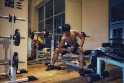 Full length of shirtless young man lifting dumbbell at gym