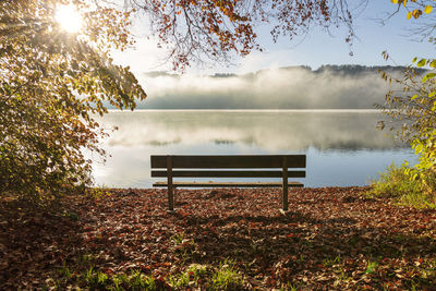 Bench by lake against sky during autumn