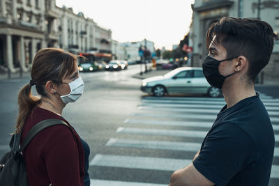 Man and woman talking and waiting at pedestrian crossing in the city center wearing the face masks