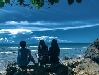 Rear view of friends sitting on rocky shore against blue sky