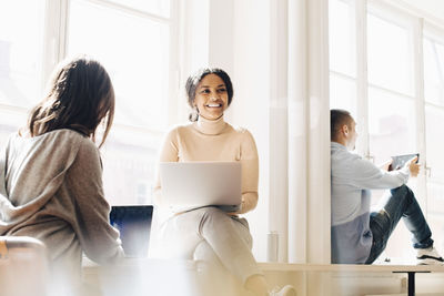 Smiling woman looking away while sitting with colleagues at desk by window in office