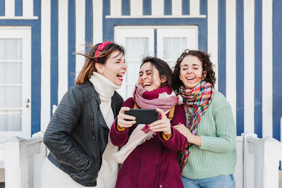 Happy women friends using mobile phone in front of colorful houses.costa nova, aveiro, portugal