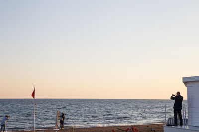 People standing by sea against clear sky during sunset