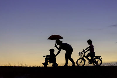 Silhouette woman with daughters riding vehicles on field against sky during sunset