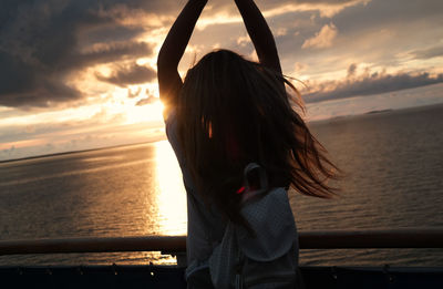 Rear view of woman standing with arms raised against sea and sky during sunset